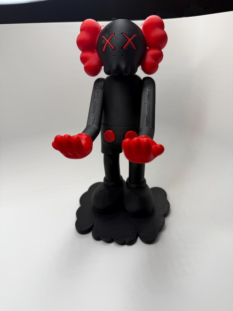 Puede incluir: Una figura negra con detalles rojos. La figura tiene una cabeza con forma de calavera con ojos tachados, manos rojas y protuberancias rojas en forma de oreja. Se apoya sobre una base negra en forma de nube. El cuerpo es negro con detalles circulares rojos.