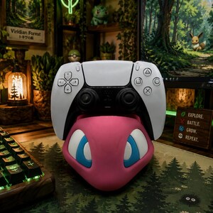 Könnte beinhalten: Ein weiß-schwarzer PlayStation 5-Controller liegt auf einem rosa Mew-förmigen Ständer mit blauen Augen. Der Controller befindet sich auf einem Schreibtisch mit einem Mauspad im Wald-Design und einer Holz-Tastatur. Ein Neon-Blatt-Schild ist im Hintergrund zu sehen.