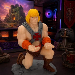 May include: A He-Man figurine kneeling on a gray rocky base, featuring blonde hair, a gray chest harness with a red cross emblem, orange wrist cuffs, and brown fur-like shorts and boots, designed to hold a mobile device in its cupped hands.