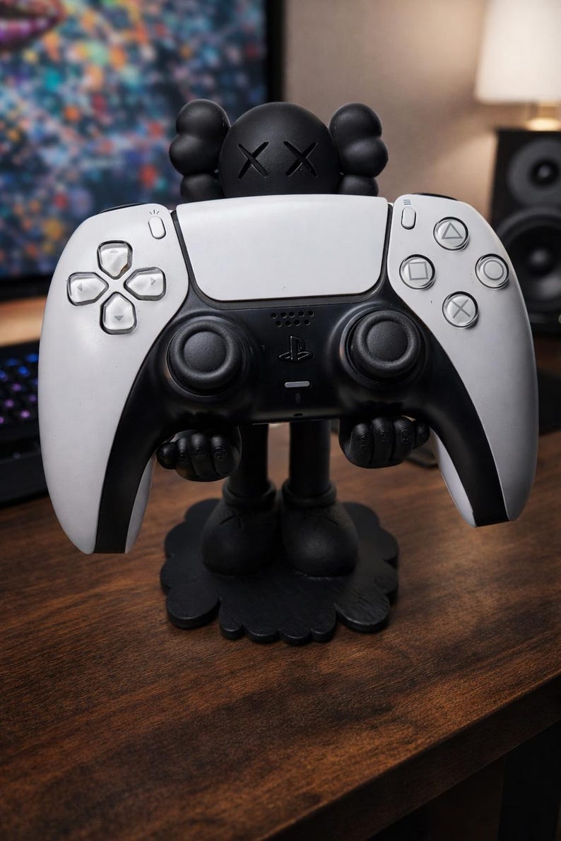 Puede incluir: Un soporte para mando de PlayStation 5 en blanco y negro con un dise&ntilde;o inspirado en KAWS. El mando es blanco con detalles en negro. El soporte es negro y descansa sobre una superficie de madera.