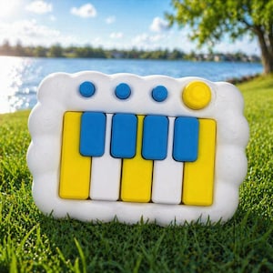 May include: A colorful toy piano with a white, cloud-shaped frame. The piano keys are yellow, white, and blue. Four blue buttons and one yellow button are on the top. The toy is set on green grass with a lake and trees in the background.