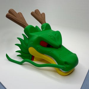 Puede incluir: Una figura de cabeza de drag&oacute;n verde con astas marrones, ojos rojos, mand&iacute;bula amarilla y dientes blancos. La criatura tiene un bigote verde largo y fino que sale del hocico y una serie de aletas verdes puntiagudas a lo largo del cuello.