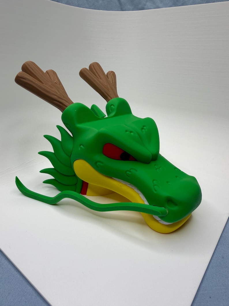 Puede incluir: Una figura de cabeza de drag&oacute;n verde con astas marrones, ojos rojos, mand&iacute;bula amarilla y dientes blancos. La criatura tiene un bigote verde largo y fino que sale del hocico y una serie de aletas verdes puntiagudas a lo largo del cuello.
