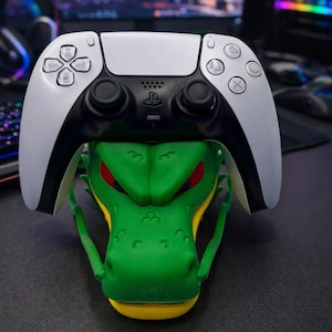 Puede incluir: Un mando blanco y negro de PlayStation 5 descansa sobre un soporte para mando con forma de dragón verde y amarillo. El soporte tiene ojos rojos. El fondo muestra una configuración informática.