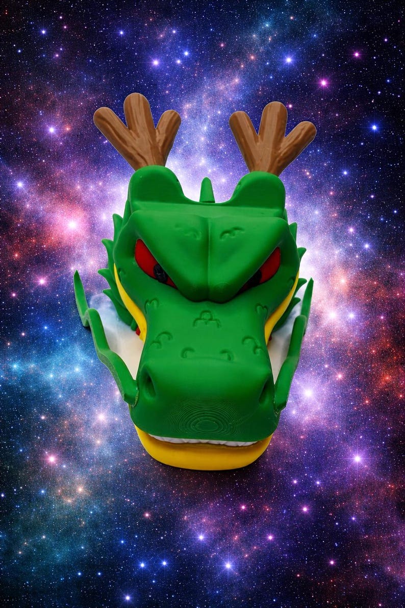 Puede incluir: Una figura de cabeza de drag&oacute;n verde con astas marrones, ojos rojos afilados y una mand&iacute;bula amarilla. La criatura tiene crestas faciales prominentes, largos bigotes laterales y una superficie texturizada como objeto escult&oacute;rico.