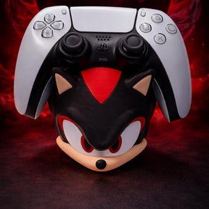 May include: A black and white video game controller resting on a sculpted stand shaped like the head of the character Shadow the Hedgehog. The stand features a black face, red markings on the forehead, red eyes, and tan ears and muzzle.