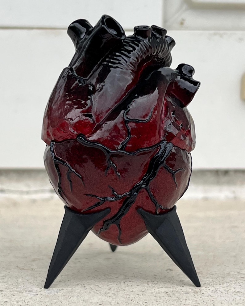May include: A sculpted anatomical heart in shades of red and black, mounted on three black, pointed legs. The heart's surface has intricate vein detailing and a glossy finish, creating a striking visual contrast.