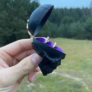 Puede incluir: Una caja de anillo de calavera negra con un interior de terciopelo morado. La caja está abierta y hay dos anillos de oro dentro.