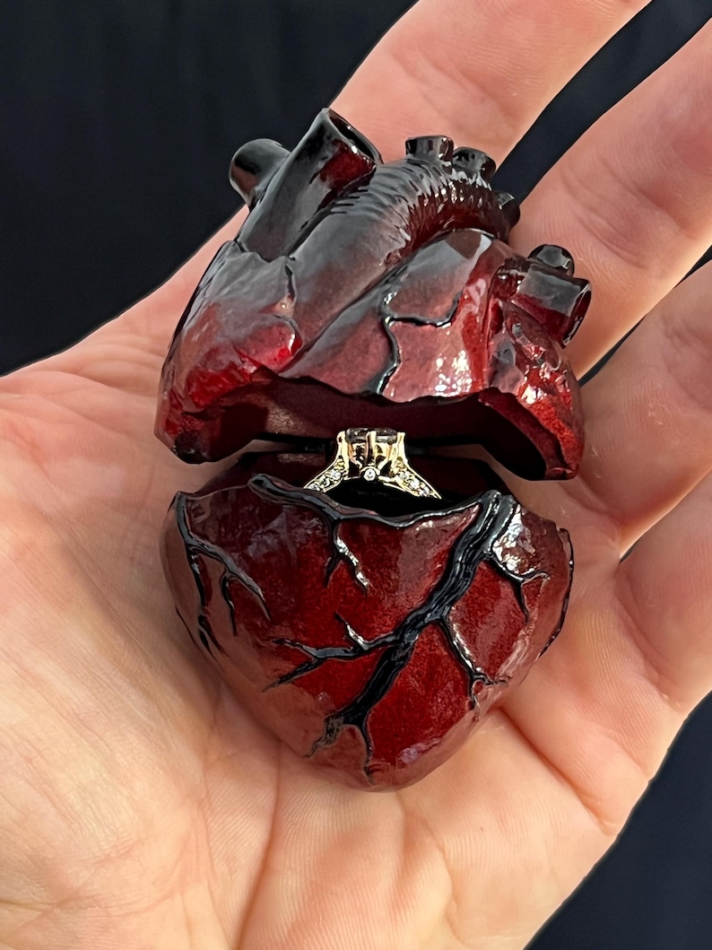 May include: A heart-shaped ring box, painted in shades of red and black, opens to reveal a diamond ring. The box is held in a hand, showcasing the intricate details of the anatomical heart design.