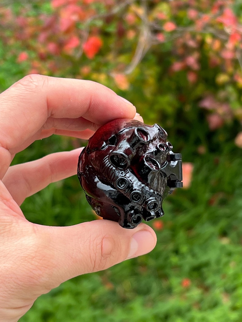 May include: A dark red and black decorative object with a complex, textured design. The object has a glossy finish and features multiple tentacle-like protrusions. The background is blurred, with green grass and colorful foliage.