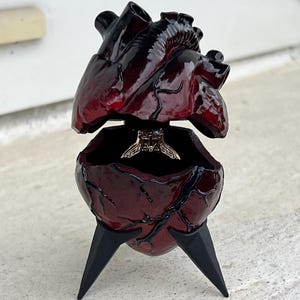 May include: A heart-shaped ring box with a dark red, glossy finish. The box is open, revealing a ring inside. The box is supported by three black, pointed legs. The top of the heart is also open.