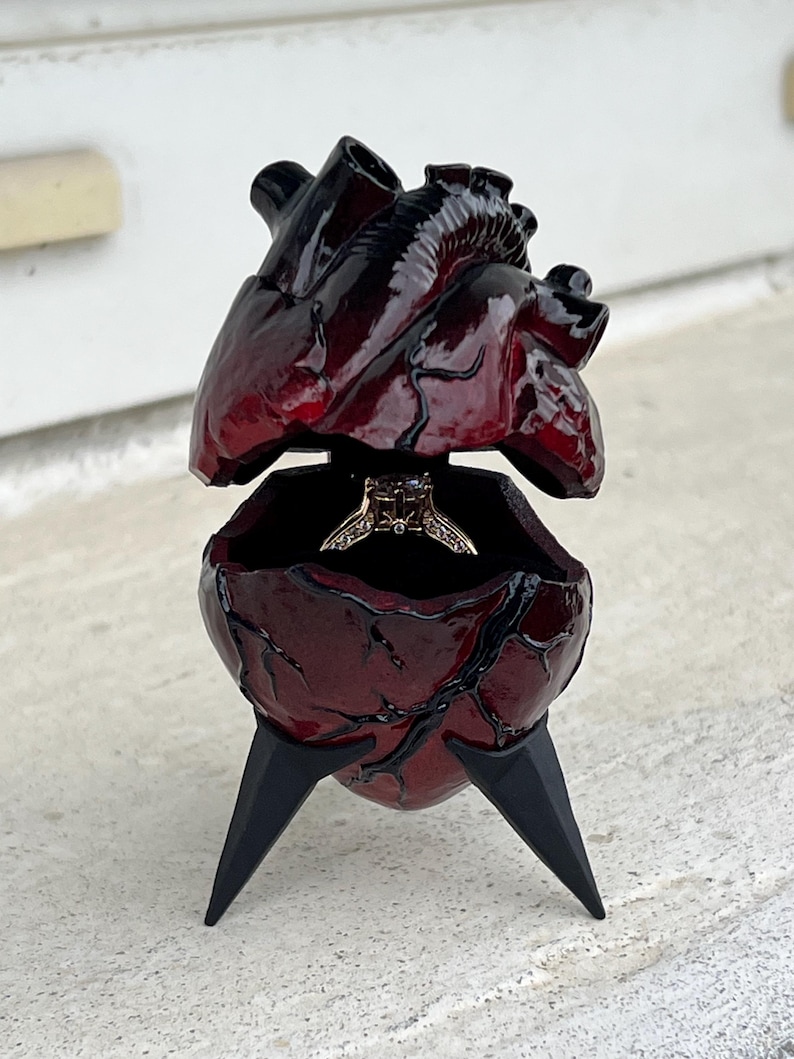 May include: A heart-shaped ring box with a dark red, glossy finish. The box is open, revealing a ring inside. The box is supported by three black, pointed legs. The top of the heart is also open.