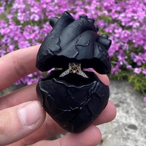Puede incluir: Un joyero negro en forma de corazón, abierto para revelar un anillo de oro con una piedra central cuadrada y diamantes. La caja se sostiene en una mano, con un fondo borroso de flores moradas.