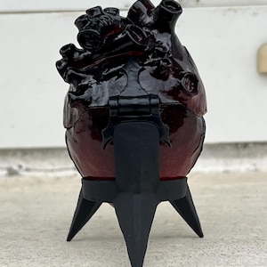 May include: A dark red, heart-shaped box with black accents and a hinged lid, mounted on a black, three-legged stand. The box has a glossy finish and detailed anatomical features.