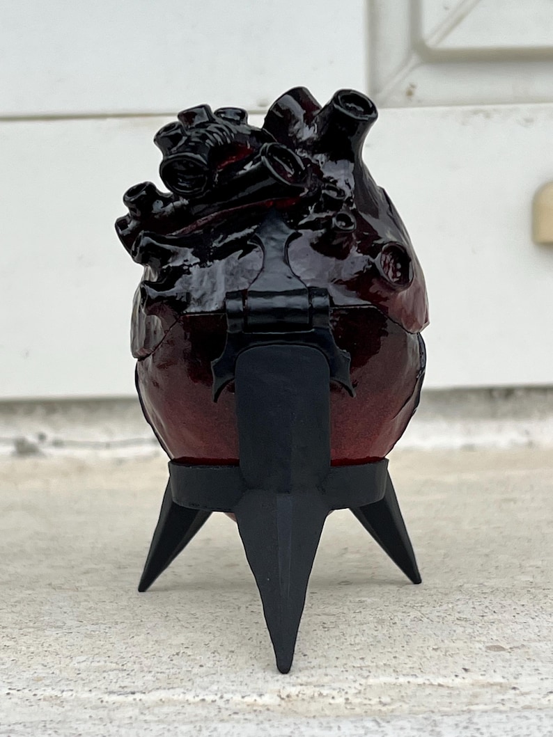 May include: A dark red, heart-shaped box with black accents and a hinged lid, mounted on a black, three-legged stand. The box has a glossy finish and detailed anatomical features.