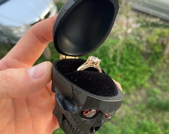 Puede incluir: Una caja de anillo de calavera negra con ojos rojos, abierta para revelar un anillo de oro con un diamante grande.