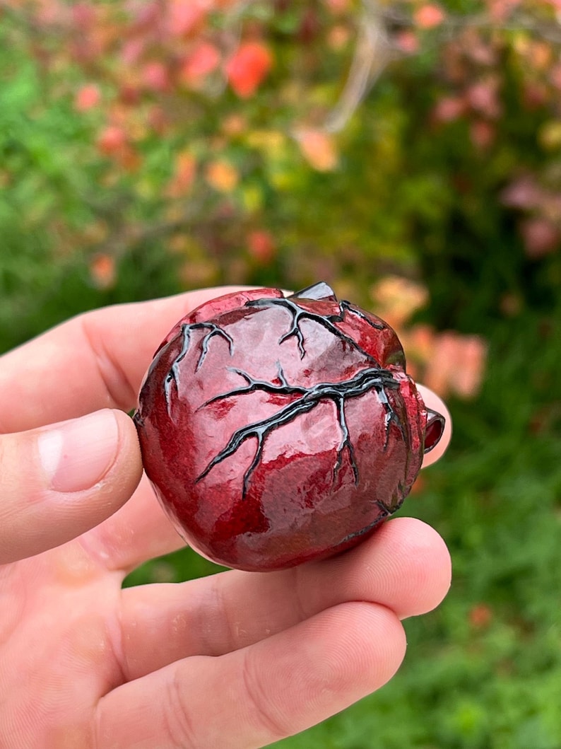 May include: A glossy, dark red heart-shaped sculpture with black vein-like details. The sculpture is held in a hand, with a blurred green and red background.