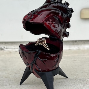 May include: A burgundy heart-shaped ring box with a hinged top, revealing a gold ring with a diamond. The box is supported by three black, pointed legs. The box has a glossy finish and a detailed, anatomical design.