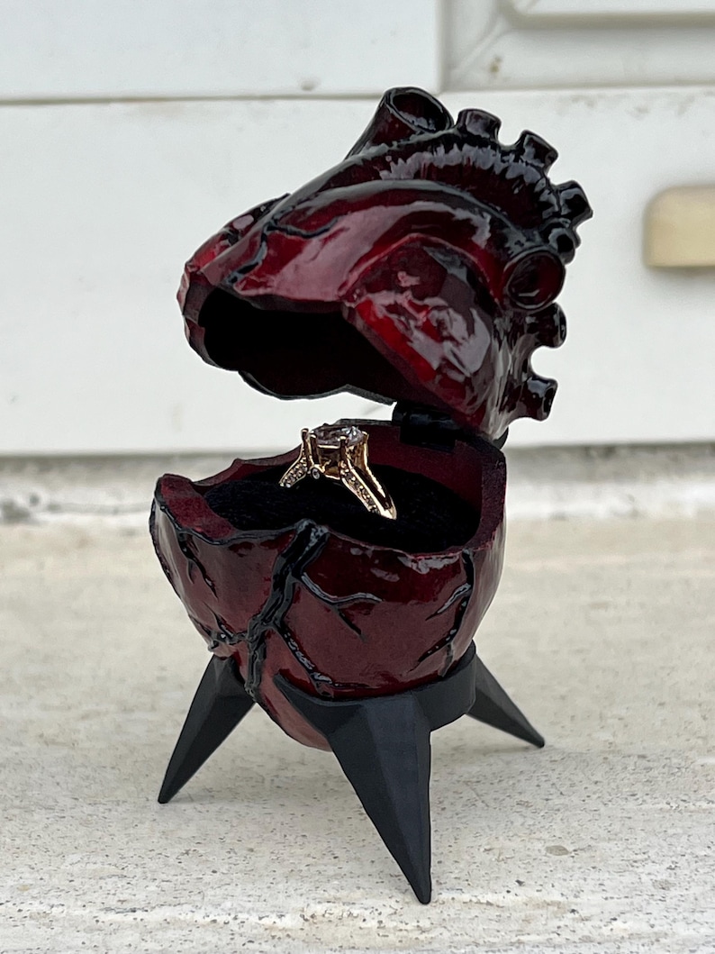May include: A burgundy heart-shaped ring box with a hinged top, revealing a gold ring with a diamond. The box is supported by three black, pointed legs. The box has a glossy finish and a detailed, anatomical design.