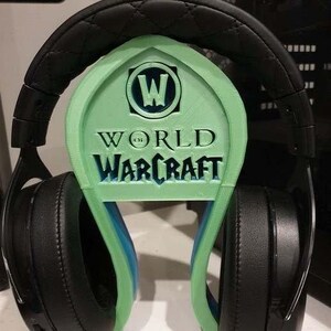May include: A green 3D printed headphone stand with the World of Warcraft logo. The stand is designed to hold a pair of black headphones.