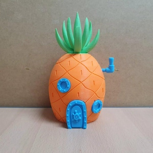 May include: A 3D printed pineapple-shaped house with a blue door and a green leafy top. The house has a blue pipe on the side and two round blue windows.