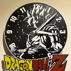 Könnte beinhalten: Eine schwarz-weiße Wanduhr mit einem Dragon Ball Z-Design. Das Zifferblatt hat weiße Ziffern und Zeiger mit einem Bild einer Figur. Das Dragon Ball Z-Logo ist in Gelb und Rot dargestellt.