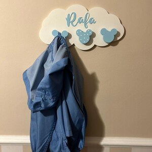 May include: A white wooden cloud-shaped wall hook with the name "Rafa" written in blue. The hook has three blue Mickey Mouse-shaped hooks with white polka dots.