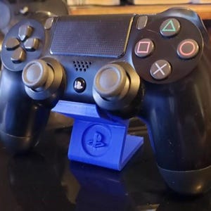 Könnte beinhalten: Ein schwarzer PlayStation 4-Controller wird auf einem blauen Ständer mit dem PlayStation-Logo präsentiert. Der Controller hat ein schwarz-weißes Design mit Knöpfen für oben, unten, links, rechts, Dreieck, Kreis, Kreuz und Quadrat.