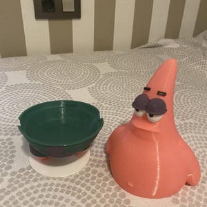 May include: A pink 3D printed figurine of Spongebob Squarepants with purple eyes and a green 3D printed bowl.