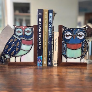 Puede incluir: Un par de sujetalibros de vidrio coloreado en forma de búho. Los búhos son azules, verdes y blancos con detalles dorados y negros. Los búhos sostienen una pila de libros con títulos como "Journey of Souls" y "Secret Language of Your Body".