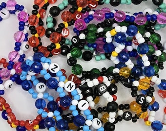 May include: A close-up of a pile of colorful beaded bracelets with letter and number charms. The bracelets are made with various colors of beads, including pink, blue, green, red, yellow, and black.
