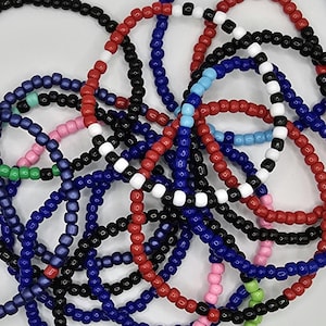 May include: A collection of colorful beaded bracelets in red, blue, black, white, pink, and green. The bracelets are made of small, round beads and are stacked on top of each other.