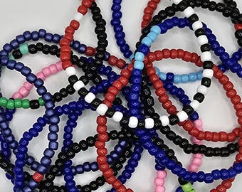 Könnte beinhalten: Eine Sammlung bunter Perlenarmbänder in Rot, Blau, Schwarz, Weiß, Rosa und Grün. Die Armbänder bestehen aus kleinen, runden Perlen und sind übereinander gestapelt.