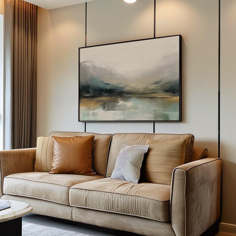 May include: A beige sofa with two brown throw pillows and a white and gray patterned throw pillow. The sofa is in front of a wall with a framed abstract landscape painting.