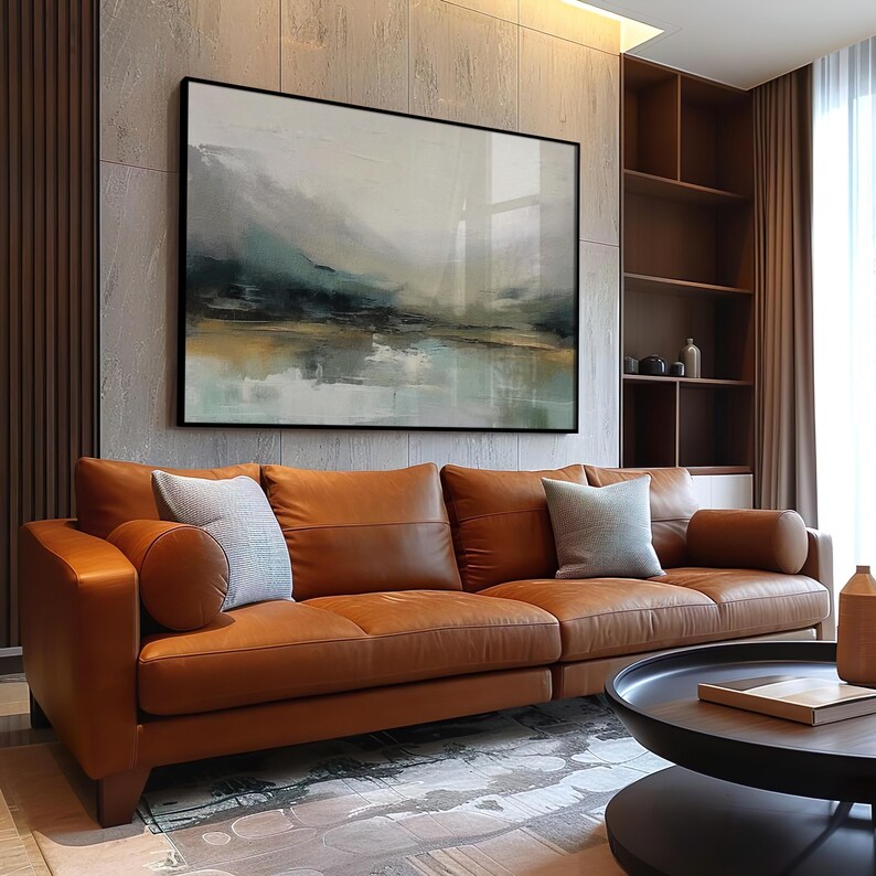 May include: A brown leather sofa with three cushions and a throw pillow. The sofa is in front of a wall with a large abstract painting of a landscape in shades of blue, green, and brown. The painting is framed in black.