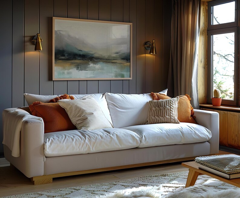 May include: A white sofa with a throw blanket and several pillows in a living room with a window and a painting on the wall.