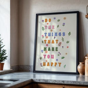 May include: A framed print with a white background and colorful text that reads "Do the things that make you happy". The text is surrounded by small floral illustrations in various colors.