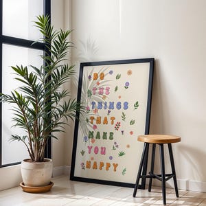 May include: A framed print with a beige background and colorful text that reads "Do the things that make you happy". The text is surrounded by small flowers and leaves. The print is hanging on a white wall next to a potted plant and a wooden stool.