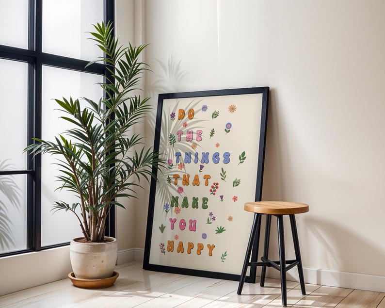 May include: A framed print with a beige background and colorful text that reads "Do the things that make you happy". The text is surrounded by small flowers and leaves. The print is hanging on a white wall next to a potted plant and a wooden stool.