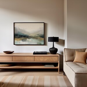 May include: A light wood media console with two drawers and a lower shelf. A black lamp with a pleated shade sits on the console next to a black book. A framed abstract landscape painting hangs on the wall above the console. A light beige sofa with a brown throw pillow is in the background.