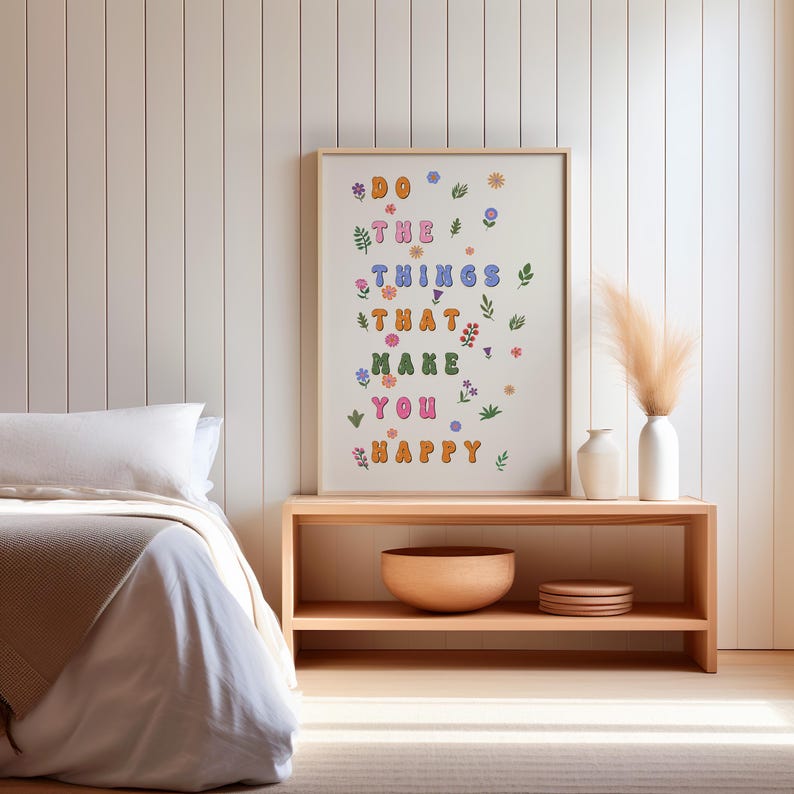 May include: A framed print with the text "Do the things that make you happy" in colorful letters with flowers and leaves surrounding the text. The print is hanging on a white wall above a wooden shelf with a bowl and decorative items.
