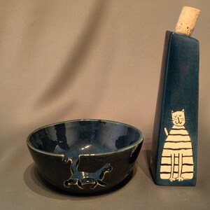 Könnte beinhalten: Ein Keramikschalen- und Flaschenset mit Katzenmotiv. Die Schale ist dunkelblau mit einem Katzensilhouetten-Design. Die Flasche hat eine dunkelblaue, rechteckige Form mit einem Korken und einer weißen Katzenillustration. Das Set ist handgefertigt.