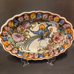 Op de afbeelding: Ovale keramische serveerschaal met een geschulpte rand. De schaal heeft een handgeschilderd ontwerp van een blauwe en gele vogel, roze en paarse bloemen en een decoratieve rand in bruin, blauw, geel en roze.