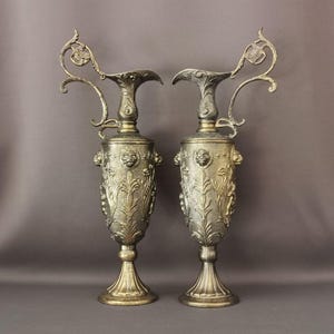 Könnte beinhalten: Zwei verzierte goldene Metallvasen mit aufwendigen Blumendesigns und Löwenkopf-Akzenten. Die Vasen haben eine hohe, schlanke Form mit einer breiten Basis und einem Ausguss an der Seite.