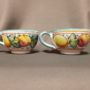 Pode incluir: Duas canecas de cer&acirc;mica branca com um desenho de frutas azul, verde, amarelo e laranja. As canecas t&ecirc;m uma forma arredondada e uma al&ccedil;a.