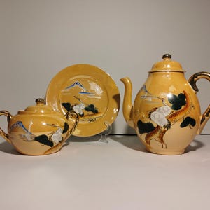 May include: A yellow tea set comprising a teapot, sugar bowl, and saucer. The set features a Japanese-inspired design with a mountain, trees, and birds painted in blue, green, and black.