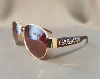 May include: Gold-rimmed aviator sunglasses with brown lenses. The sunglasses have a black frame with the text "DOLCE & GABBANA" on one arm and "GABBANA" with rhinestones on the other arm.