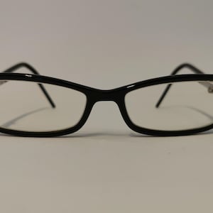 Op de afbeelding: Zwarte bril met rechthoekige glazen. De bril heeft een glanzende afwerking en een klassiek ontwerp. De pootjes zijn dun en lopen door tot de oren. De bril staat tegen een effen witte achtergrond.