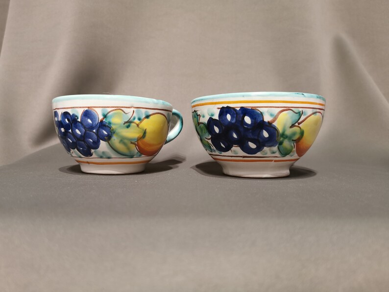 Pode incluir: Duas tigelas de cer&acirc;mica branca com um desenho de frutas azul, verde e amarelo. As tigelas s&atilde;o decoradas com uvas, folhas e uma p&ecirc;ra.