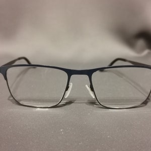Może przedstawiać: Okulary metalowe w kolorze czarnym z przezroczystymi szkłami. Okulary mają prostokątne oprawki i leżą na szarej powierzchni.